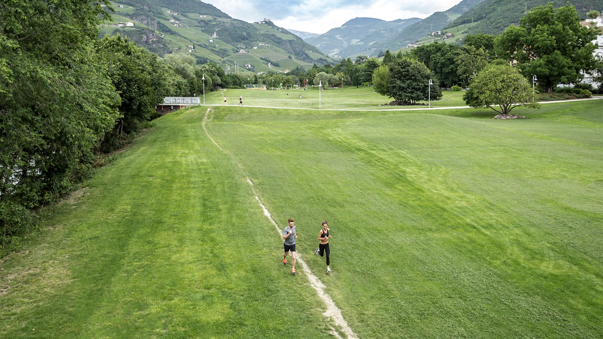 Two sports enthusiasts are running on the green meadows along the Talvera river. 