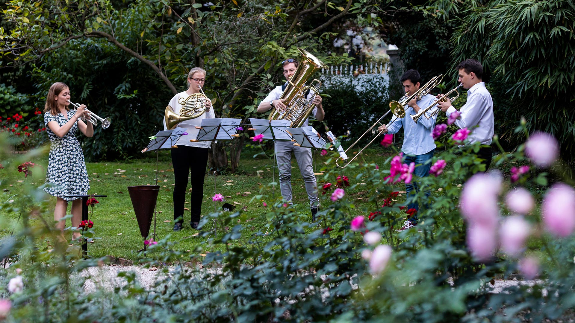 Eine Gruppe von fünf jungen Leuten, die an einem Sommernachmittag auf einer Bozner Wiese verschiedene Blasinstrumente spielen.