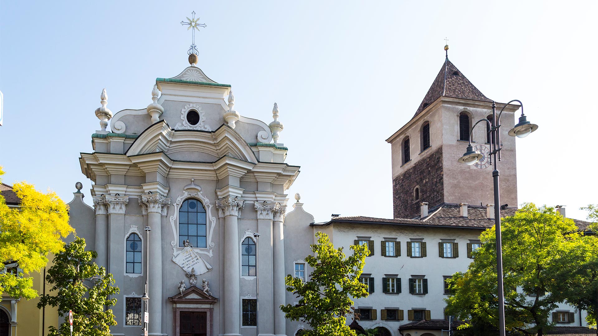 Ein Blick auf eine traditionelle Kirche in Bozen an einem sonnigen Tag. Daneben steht ein alter Glockenturm, der die Uhrzeit 4 Uhr nachmittags anzeigt.