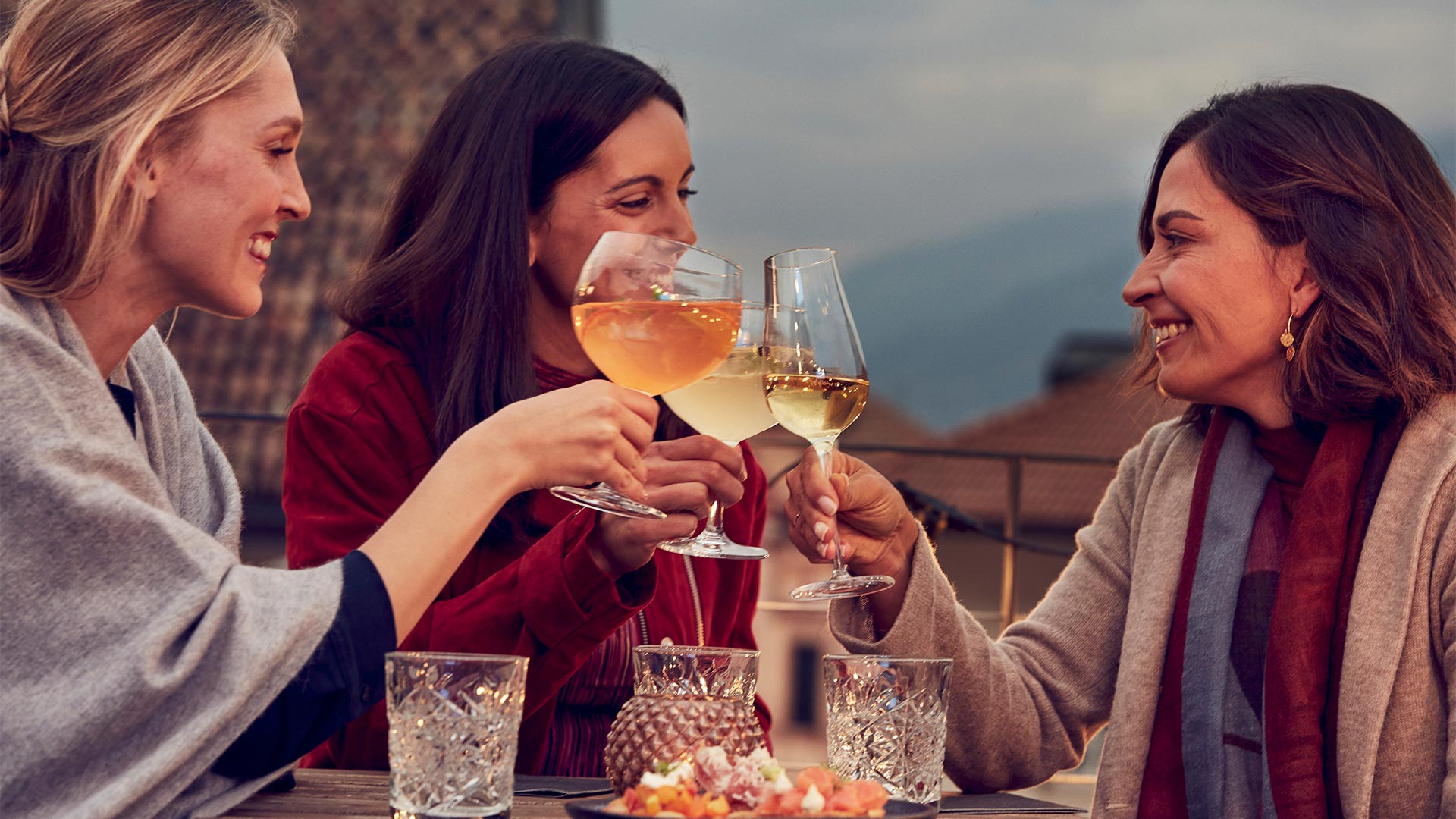 Di sera tre amiche sedute su un tavolo di un ristorante bolzanino all'aperto fanno un brindisi con dei bicchieri di vino bianco.