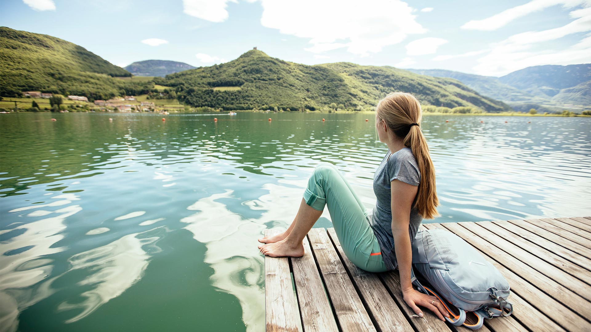 Ein Mädchen sitzt auf dem Steg des Kalterer Sees und schaut auf das Dorf auf der anderen Seite des Sees und die vorbeifahrenden Boote.