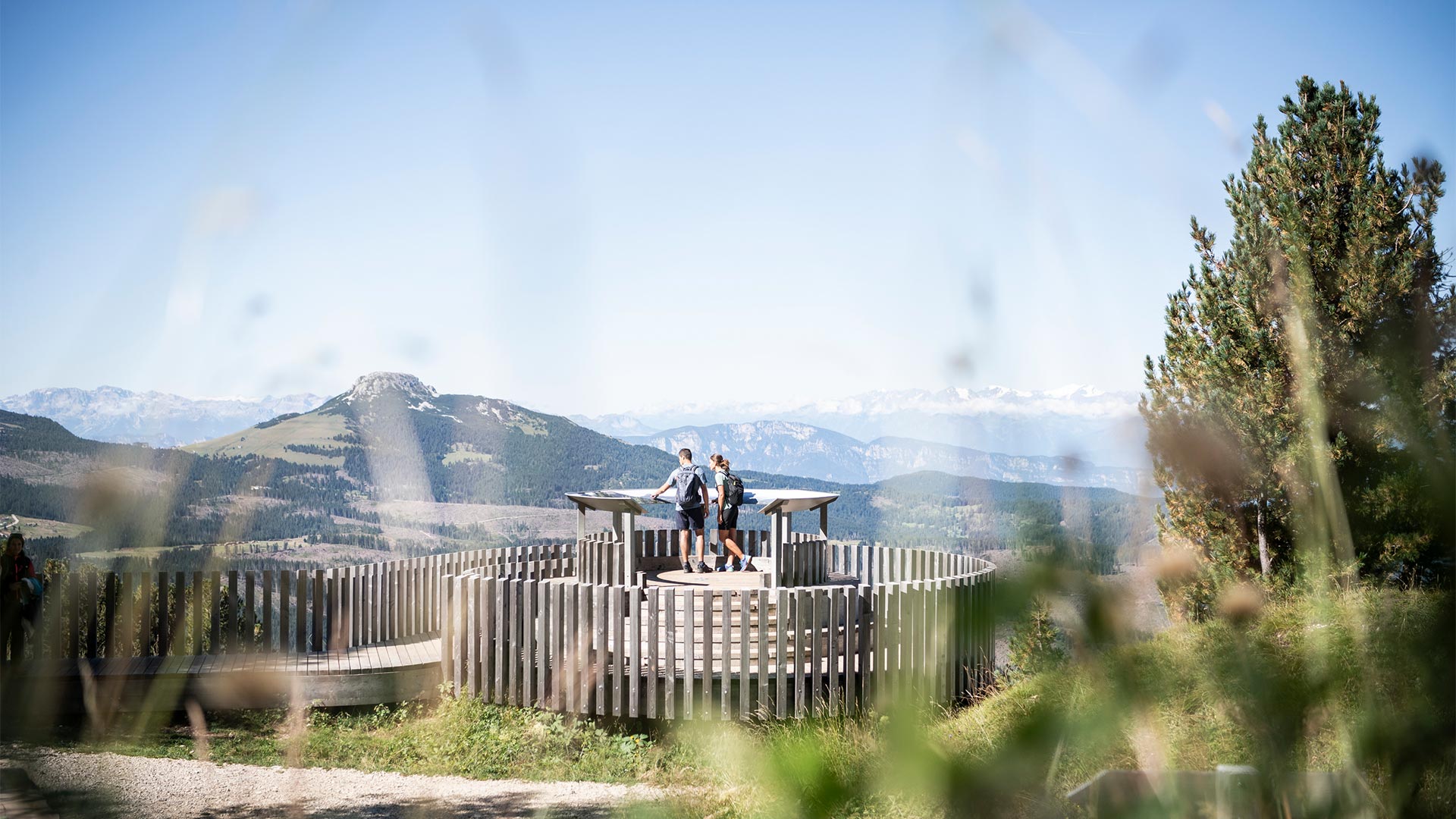 Two hikers stand at a point of interest on the Bolzano peaks and read information about the history and geography of the landscape.