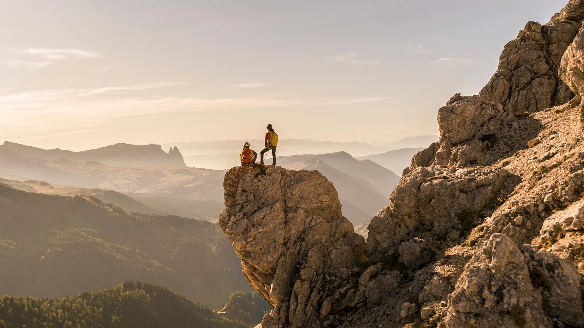 Two climbers on a cliff enjoy a panoramic view of Bolzano's natural landscapes and are inspired by their beauty.
