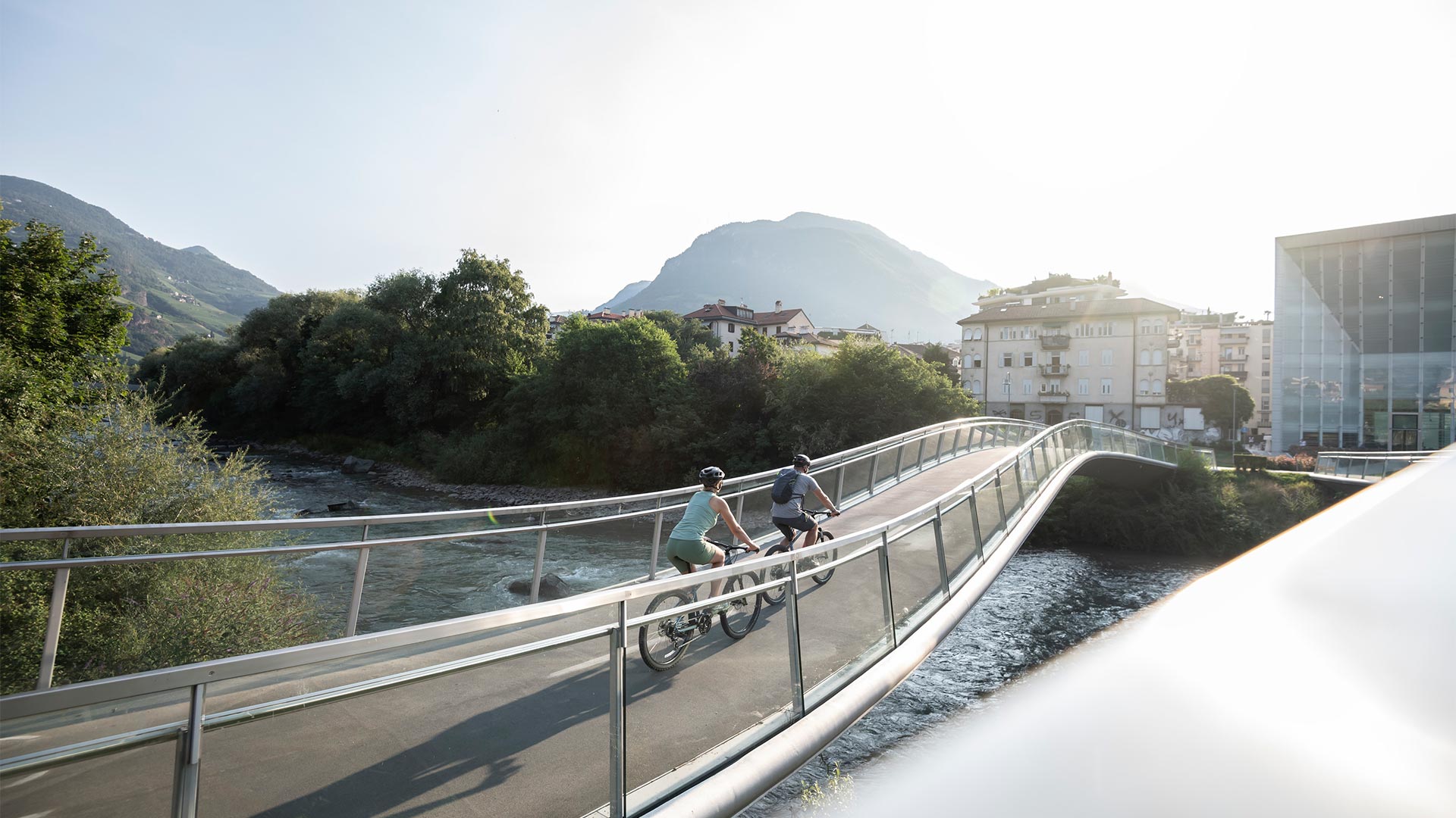 Two young people on mountain bikes cross the Museion bridge on a sunny day.