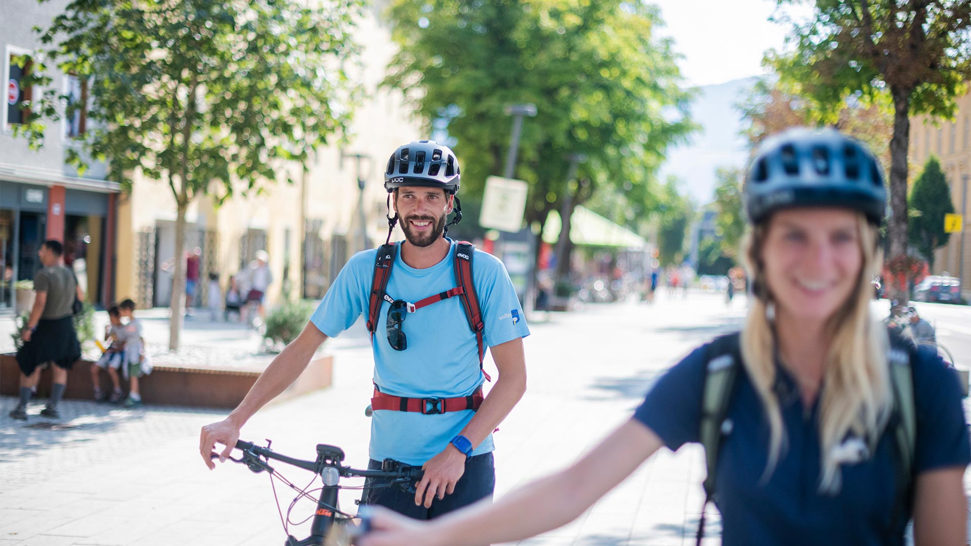 In primo piano un ciclista vestito di azzurro cammina lungo le vie di Bolzano sorridendo con la sua mountain bike in mano.