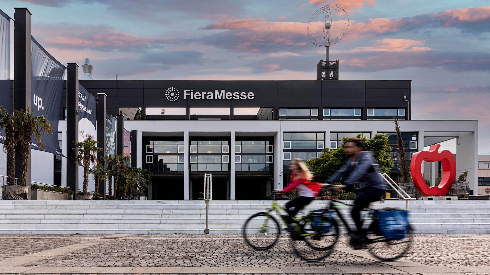 In front of the Bolzano trade fair, two friends are cycling along the cycle path. 