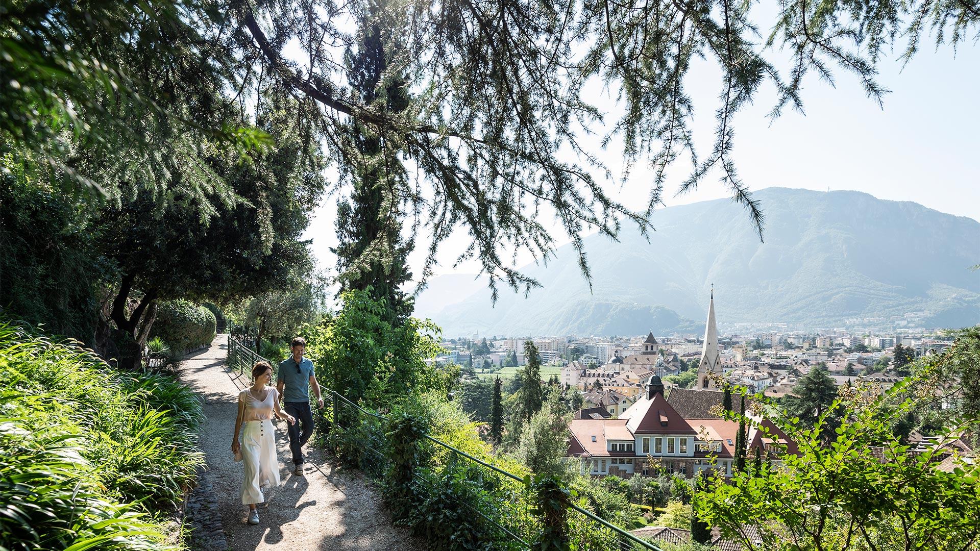 An einem schönen sonnigen Tag schlendert ein verliebtes Paar entlang der Oswaldpromenade mit Blick auf das Wohngebiet von Bozen.