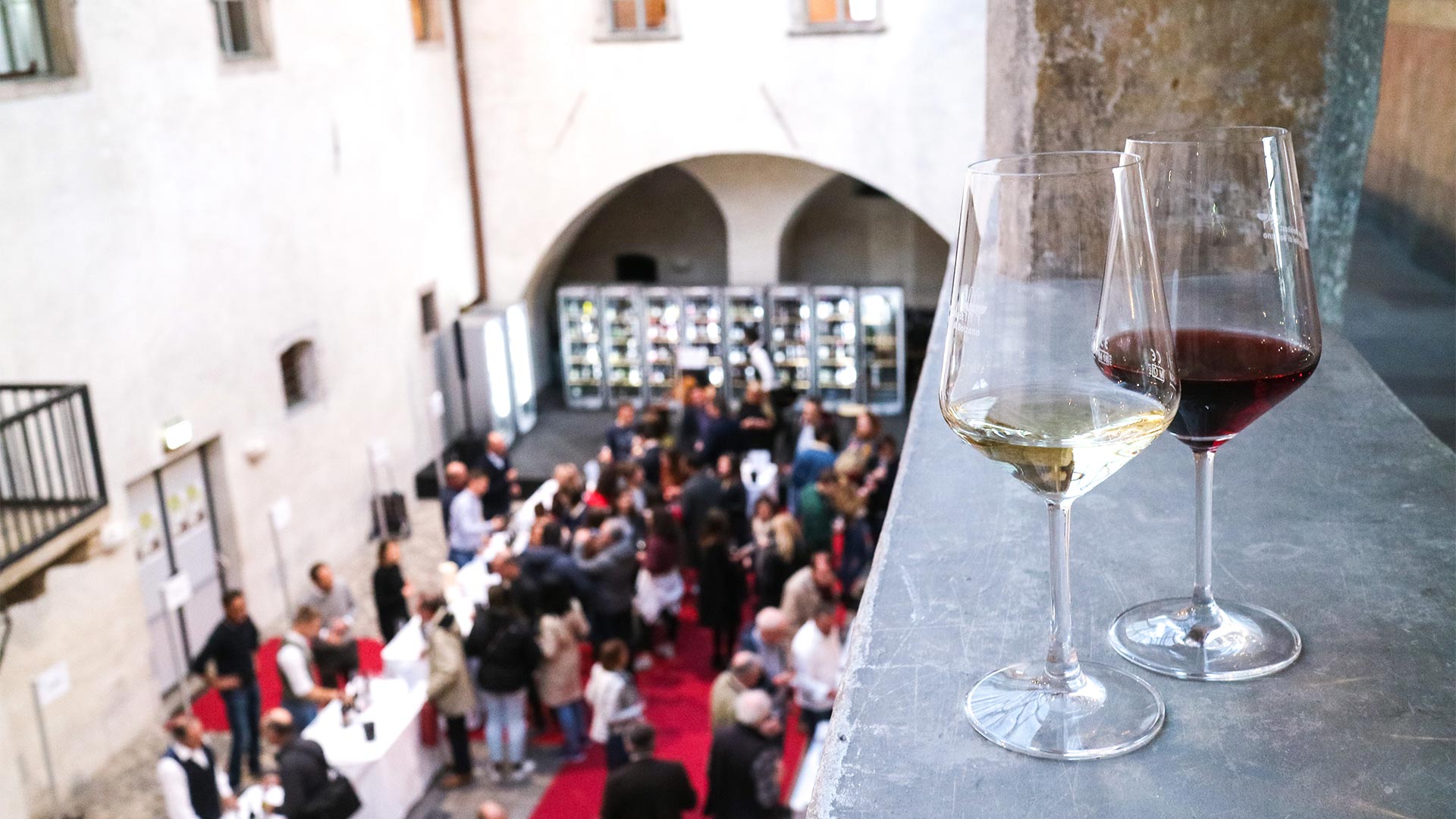 In the foreground two glasses of red wine resting on the window of a building under which there is a crowd of tourists and citizens attending a food and wine event.