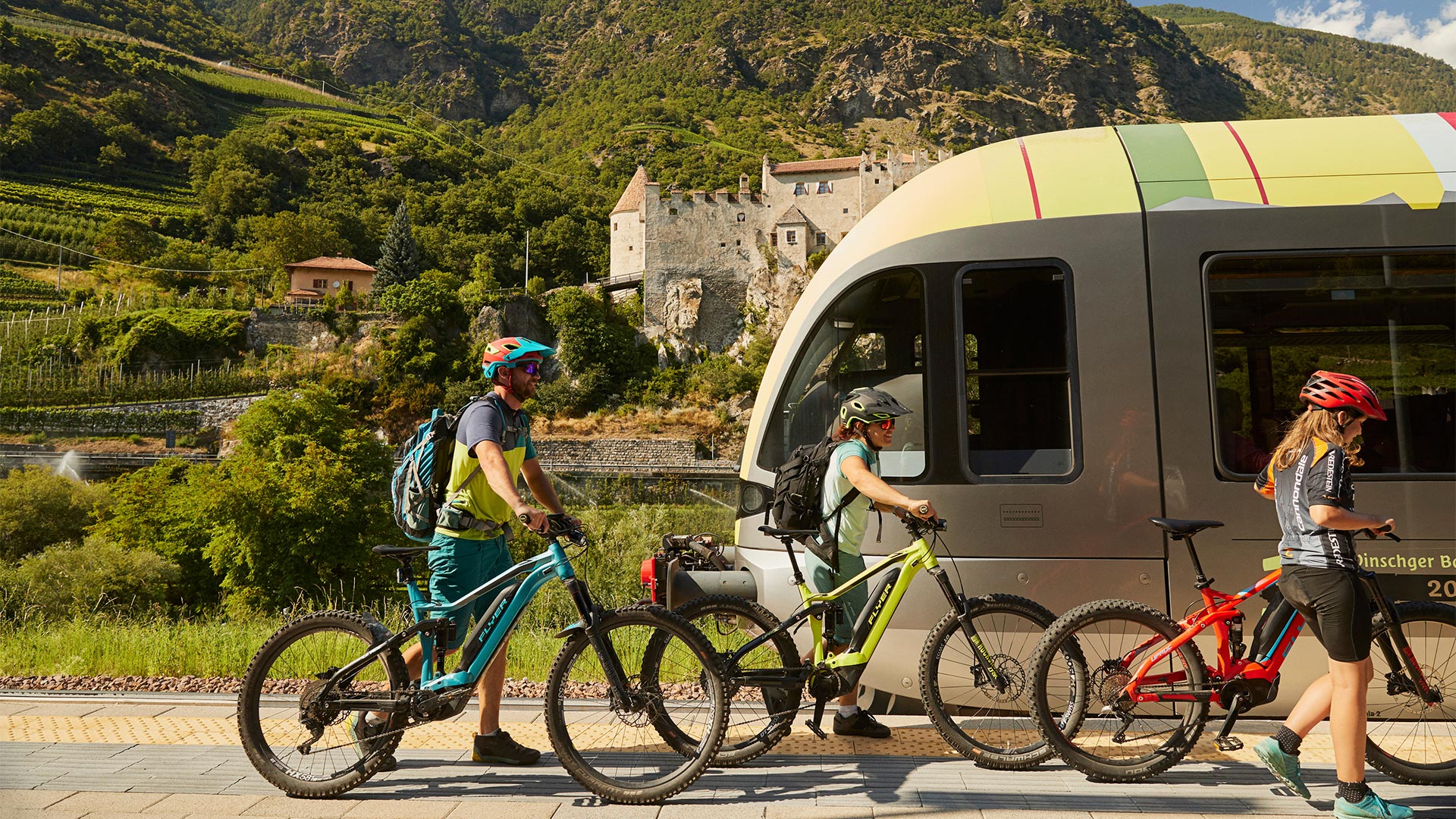 La famiglia in primo piano si muove lungo il treno con le bici trainate a mano. Sullo sfondo si vede il paesaggio di un paese con un antico castello.