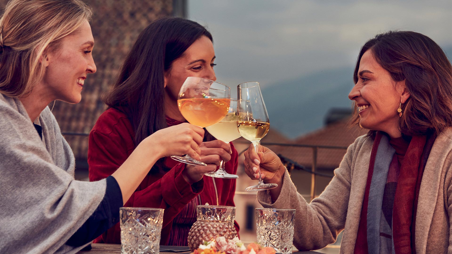 In the evening, three friends sitting at a table in an open-air restaurant make a toast with glasses of white wine.