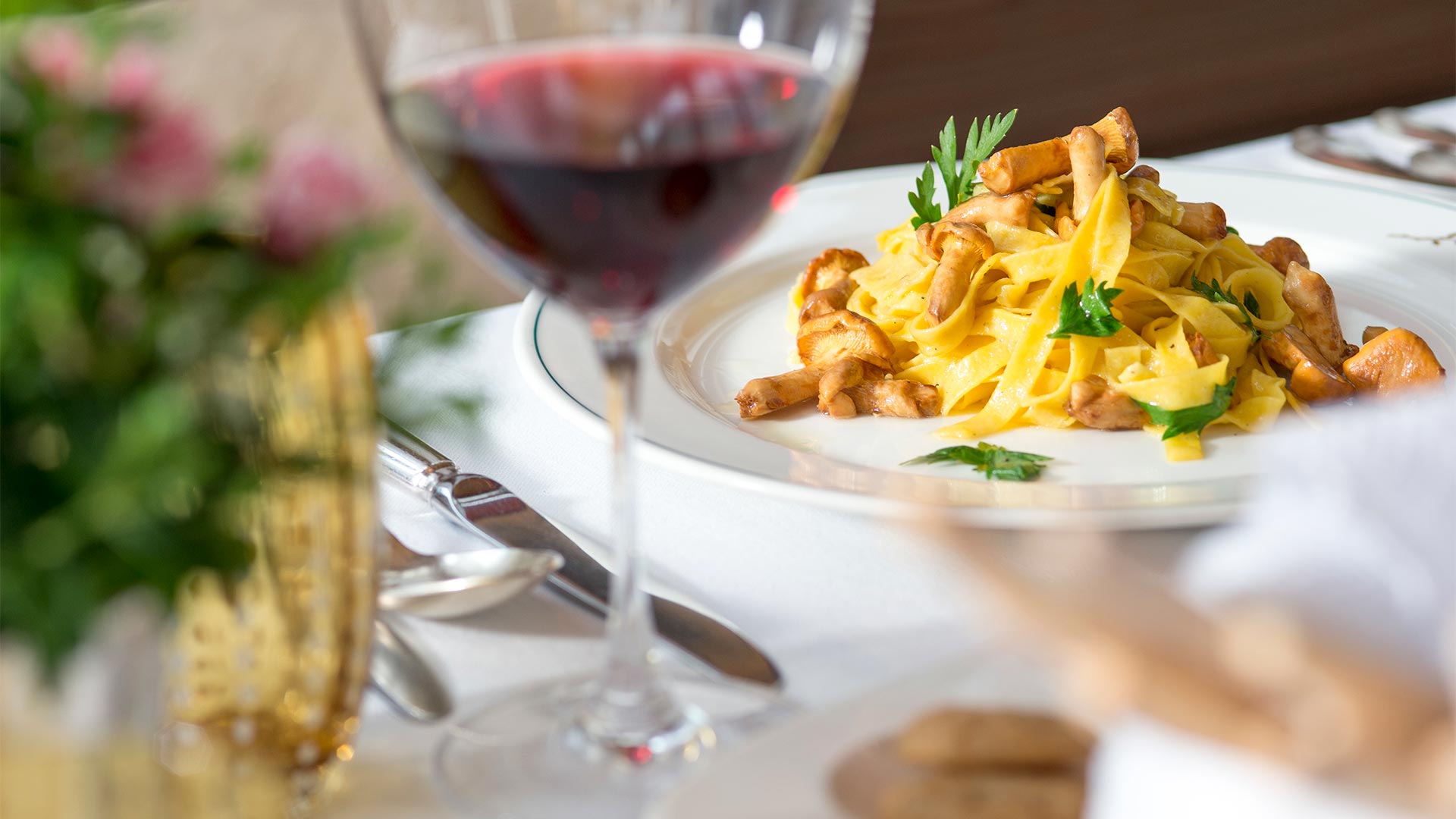 Auf einem Tisch in einem Restaurant mit weißem Tischtuch steht ein Teller mit Tagliatelle mit Fleischsauce, dazu ein Glas Rotwein.