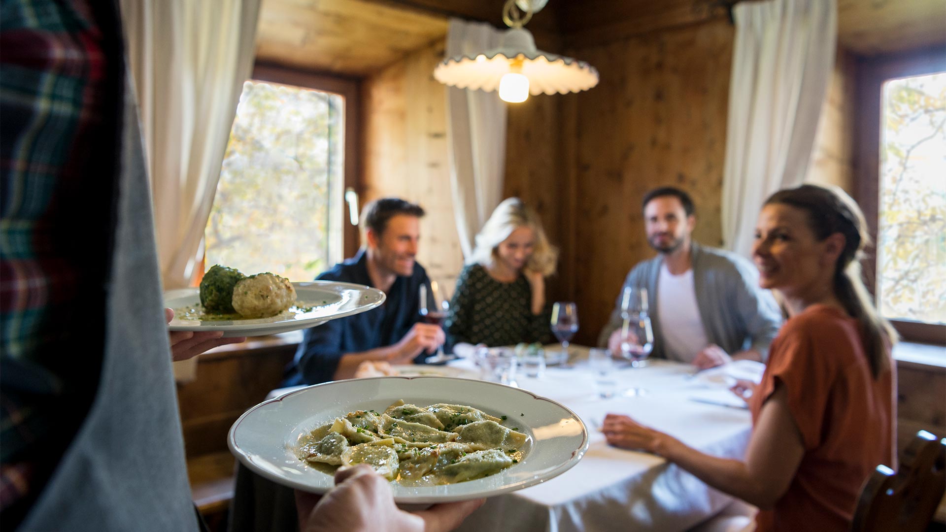 Ad un gruppo di amici seduto sul tavolo di un ristorante a Bolzano, il cameriere sta per servire i piatti da loro ordinati. I bicchieri di vino sono già stati serviti.