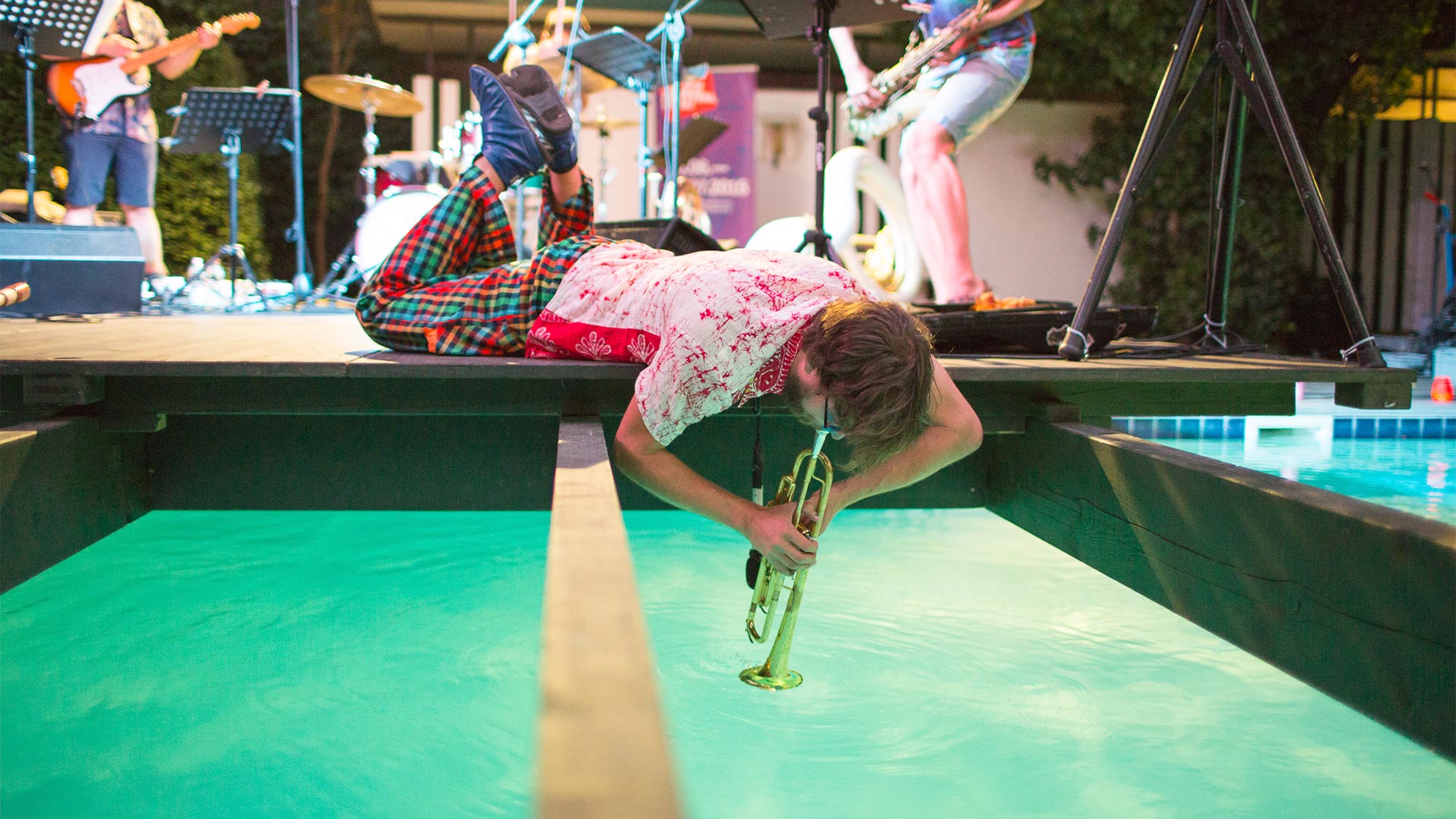 In a summer music concert a trumpeter is lying on his stomach on the edge of a platform above the water and playing his trumpet.