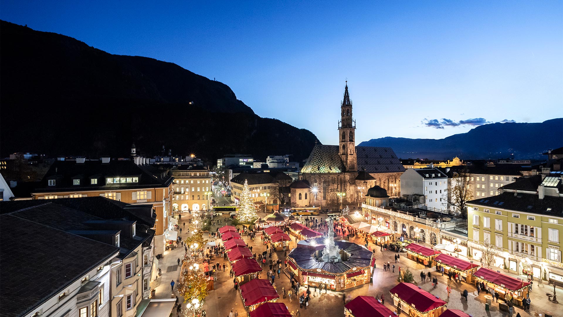 A bird's eye view of a traditional event on Piazza Walther on a winter's evening, where Christmas markets occupy the surface and crowds of shoppers are intent on shopping. 