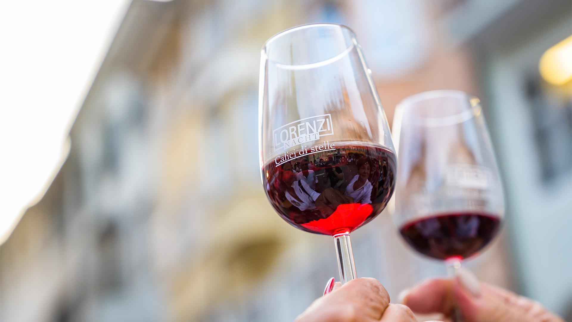 Two glasses of red wine in the foreground held with outstretched arms between two people in the middle of an event.