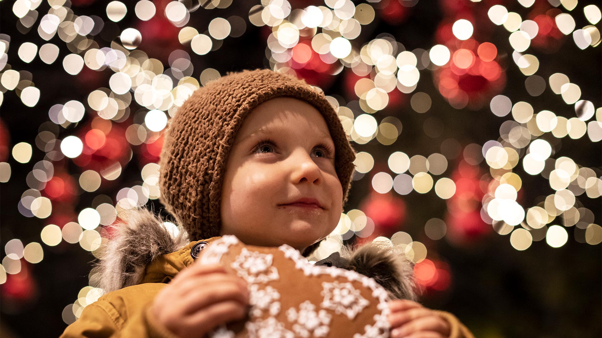 Against a backdrop of lights, in the foreground a child eats a sweet in a Christmas atmosphere.