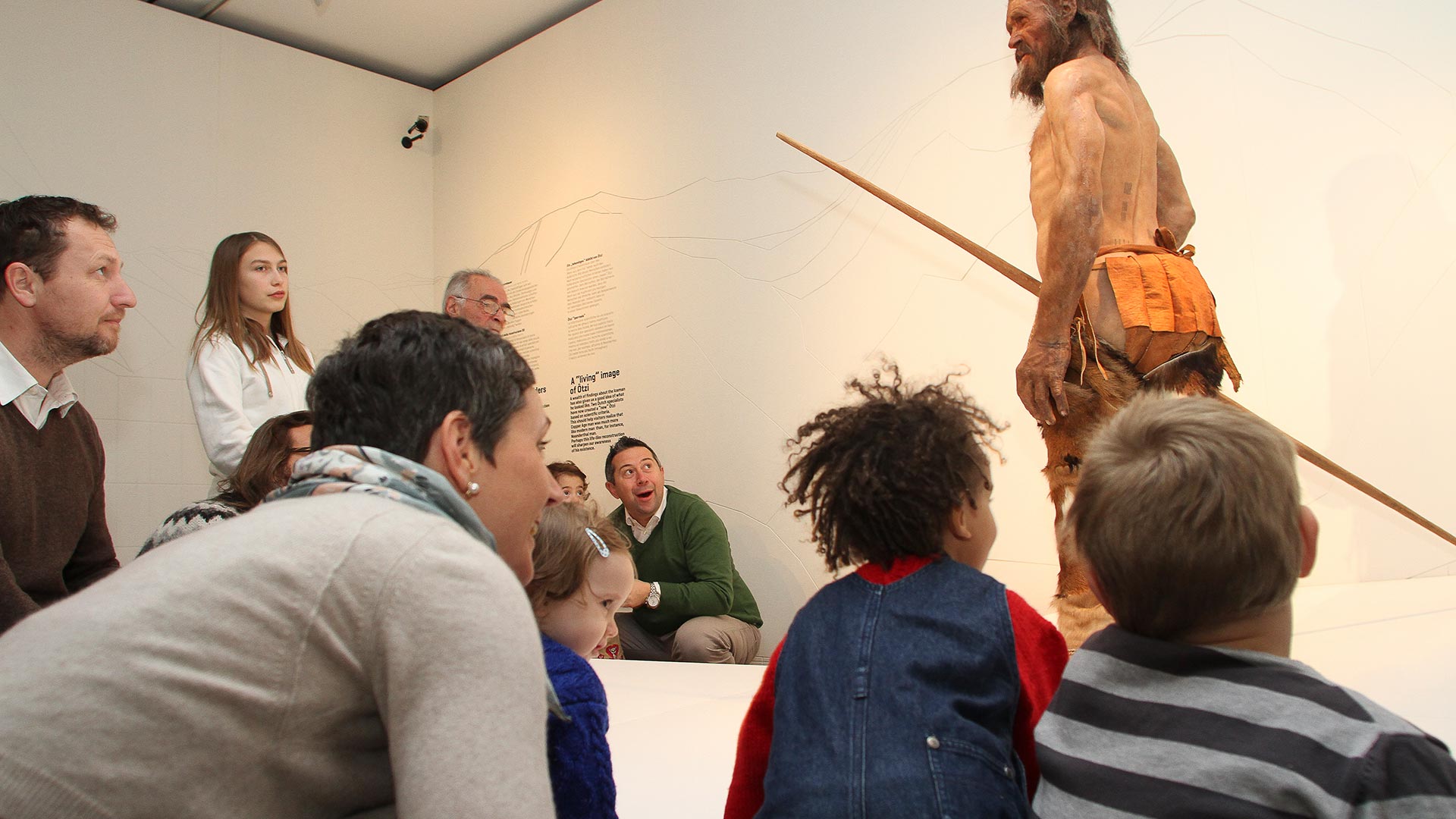 Nel museo di Ötzi delle famiglie di giovani e adulti sono sedute in cerchio attorno a una guida e ammirano la riproduzione su scala naturale dell'uomo delle nevi. 