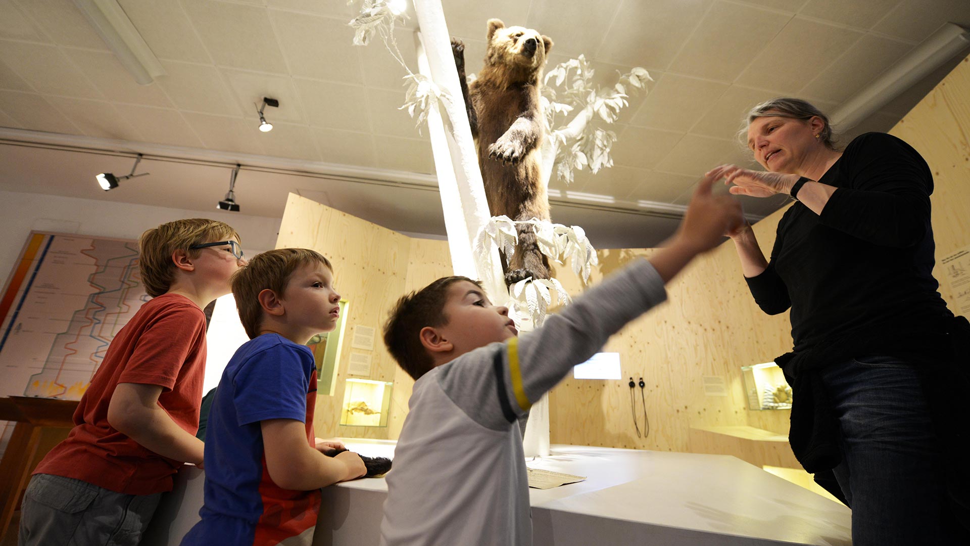 Eine schwarz gekleidete Führung zeigt drei blonden Kindern, was es im Museum zu sehen gibt.