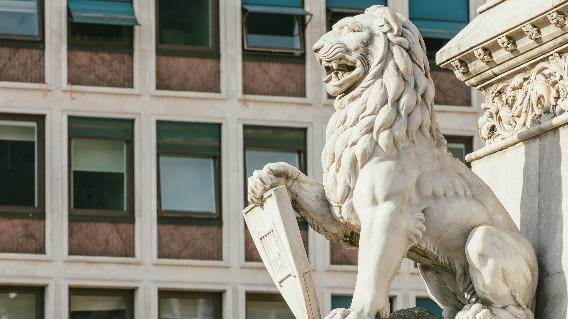 A stone sculpture of a lion holding a shield under its paw stands next to a building.