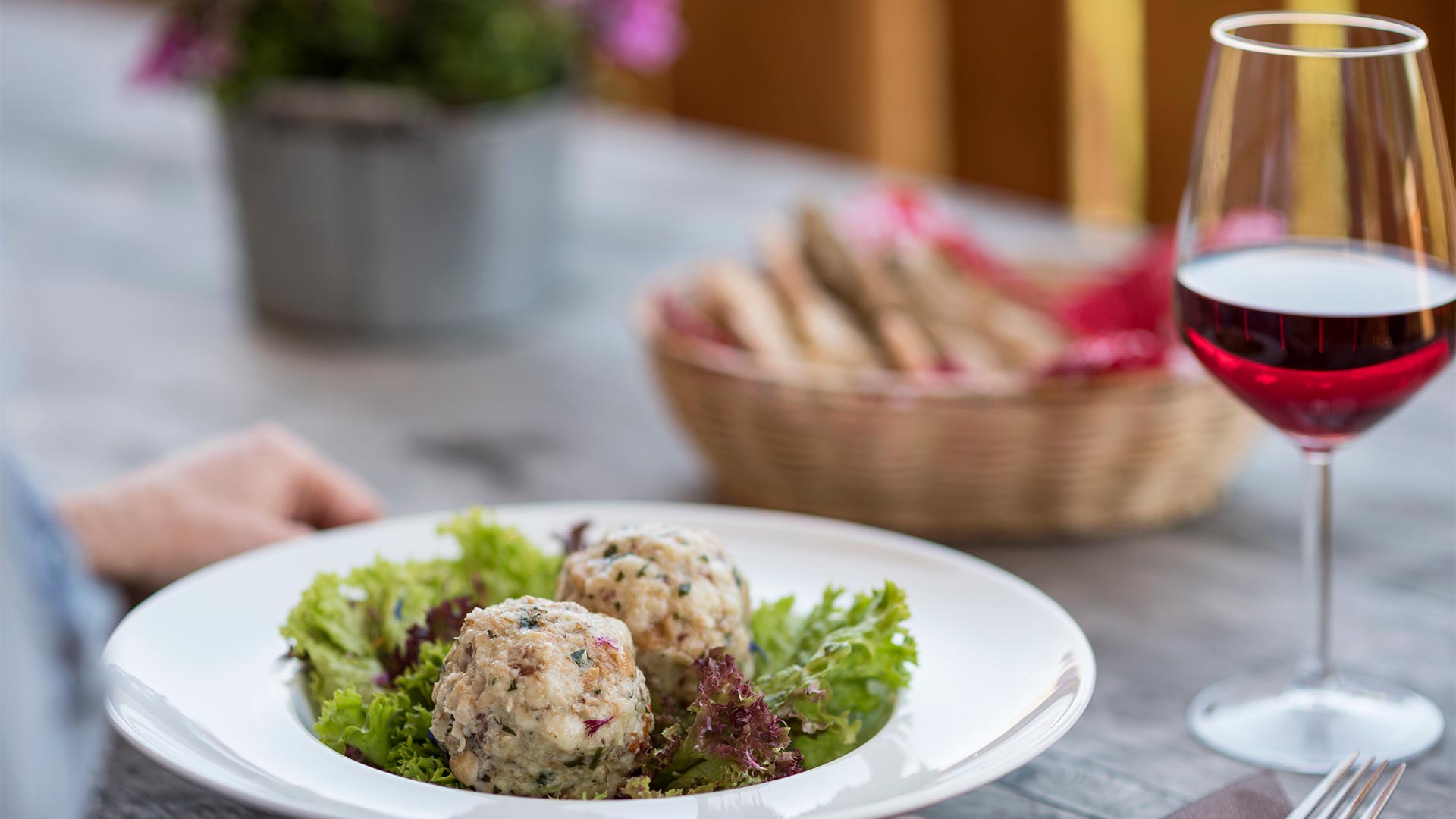 Auf dem Tisch steht ein typisches Tiroler Gericht mit Knödeln und Salat als Beilage, dazu ein Korb mit Weißbrot und ein Glas Rotwein.
