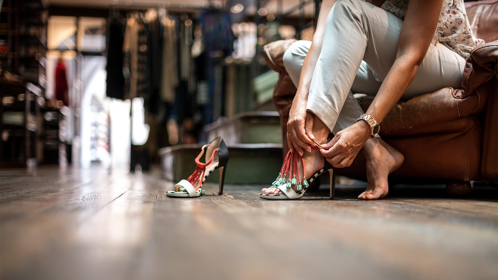 In primo piano un paio di scarpe da donna coi tacchi poggiati sul pavimento di un negozio di abbigliamento e una donna seduta accanto su una sedia intenta a provarle.