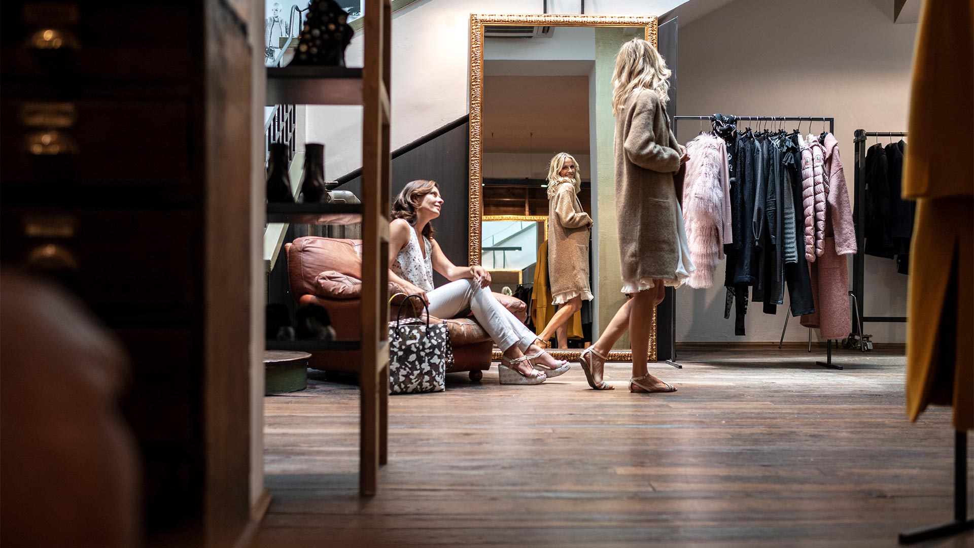 A girl is trying on a coat and is looking at herself in the mirror while her friend is sitting in a small armchair watching her.