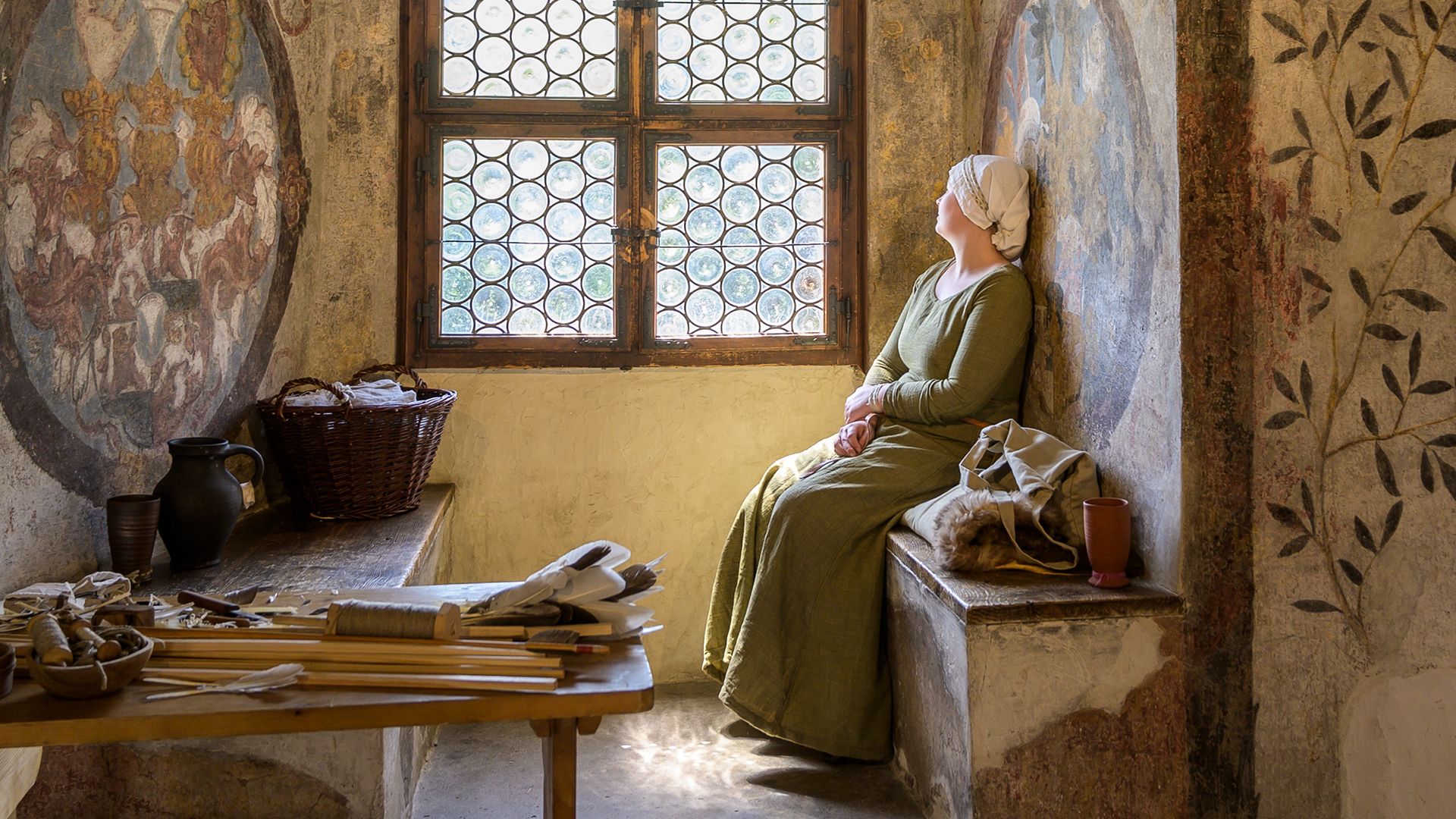 A woman, dressed in a traditional way, is sitting on a wooden bench inside an old room and looks out of the window. In front of her are domestic tools resting on a wooden table. 