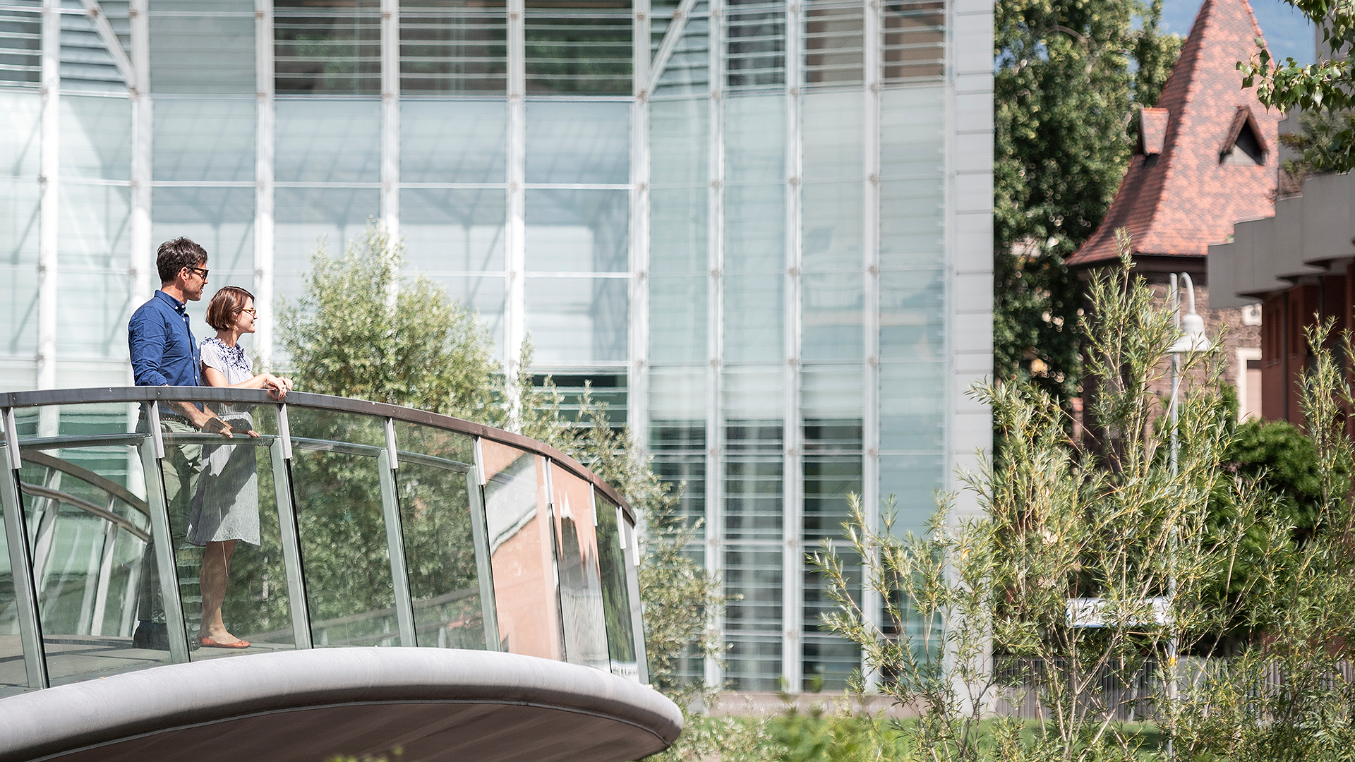 A couple of visitors stop on the Museion bridge and are inspired by the beauty of the river and the green meadows of the Talvera in Bolzano.