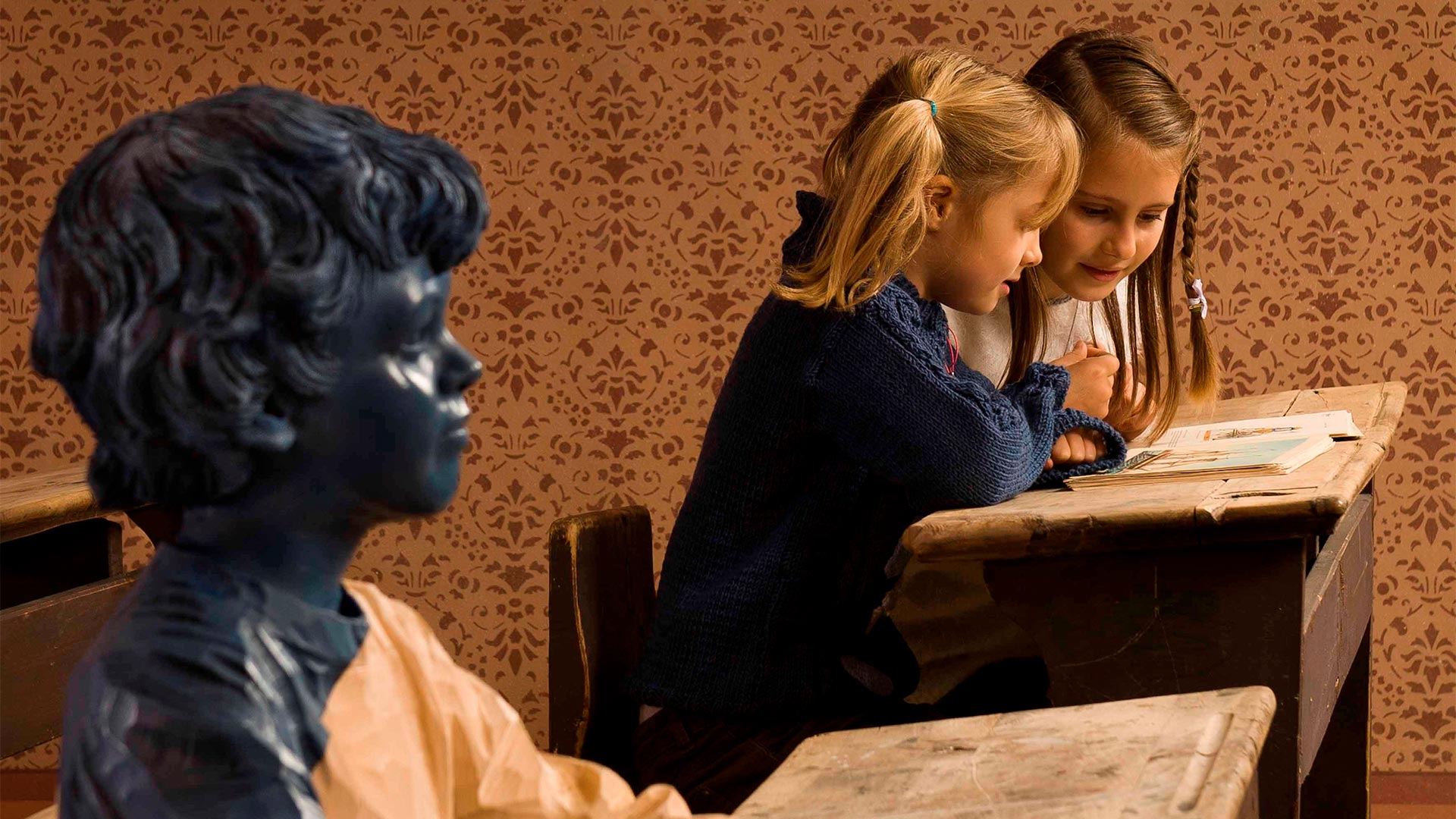 In a museum room, two sisters are leafing through leaflets with pictures of history. Next to them is a bronze sculpture of a child. 